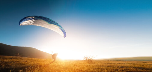 A paraglider glides along the ground at sunset with mountains in the background. Panoramic shot banner for paragliding in warm colors. Glare from the sun in the frame - obrazy, fototapety, plakaty