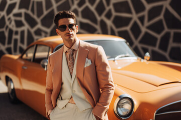 Stylish man in a peach suit posing beside a classic car. Peach Fuzz color of the 2024. Vintage fashion. Suitable for fashion brand advertising, banner, or promotions
