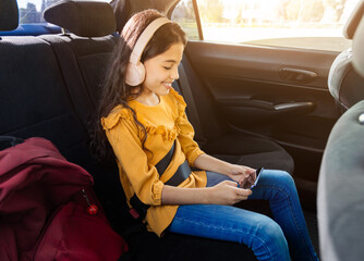 Smiling girl in headphones using phone in car sitting on backseat