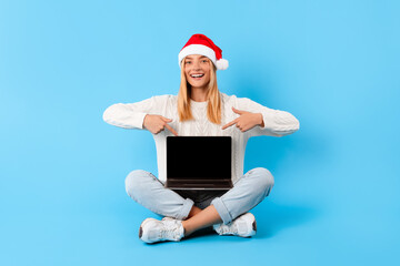 Cheerful lady in Santa hat pointing at laptop screen, blue background