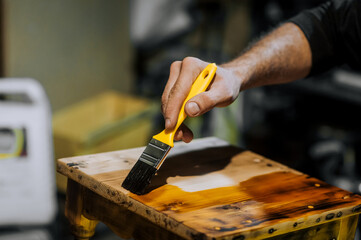 A man, a professional worker, varnishes furniture with a brush, painting a wooden chair with varnish in the workshop. Photography, handcraft work concept.