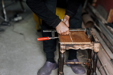 An adult male professional worker, woodworker, carpenter glues a chair, repairs, restores furniture indoors, workshop, clamping wooden products with a clamp. Photography, work concept, portrait.