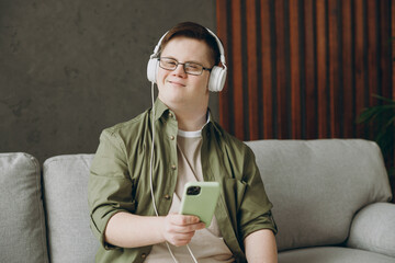 Young man with down syndrome wear clothes headphones listen music use mobile cell phone sits on...