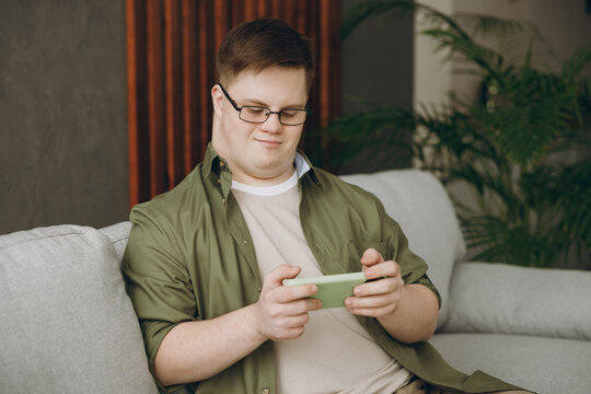 Young man with down syndrome in casual clothes use play racing app on mobile cell phone sit on grey sofa couch stay at home flat rest spend free time in living room. Genetic disease world day concept © ViDi Studio