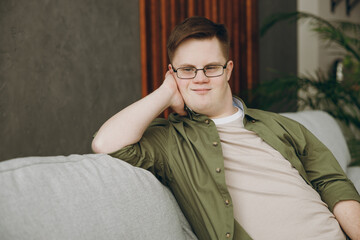 Side view young minded man with down syndrome wear glasses casual clothes look aside sits on grey...