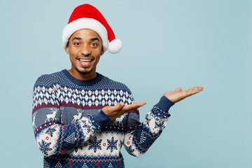 Young smiling fun man wear knitted sweater Santa hat posing point hands arms aside on area mockup isolated on plain pastel blue background. Happy New Year 2024 celebration Christmas holiday concept.