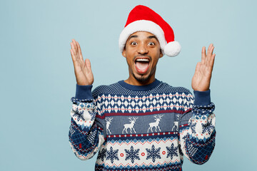 Young shocked surprised cool man wears knitted sweater Santa hat posing look camera spreading hands...