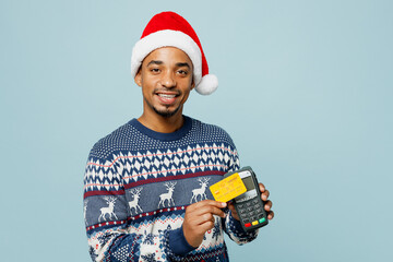 Young man wear sweater Santa hat posing hold wireless modern bank payment terminal process acquire credit card isolated on plain pastel blue background. Happy New Year 2024 Christmas holiday concept.
