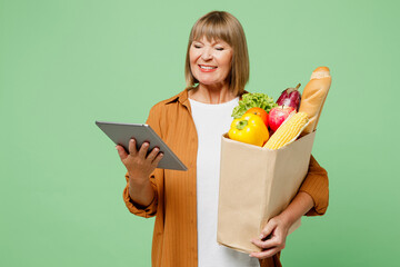 Elderly smiling happy woman wear brown shirt casual clothes hold shopping paper bag with food products hold digital tablet pc computer isolated on plain green background. Delivery service from shop.