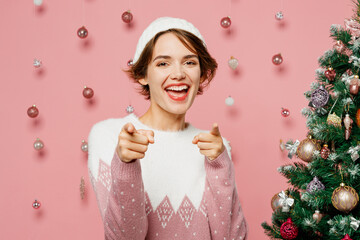Merry young woman wears white sweater hat posing point index finger camera on you motivating encourage isolated on plain pastel pink background. Happy New Year celebration Christmas holiday concept.