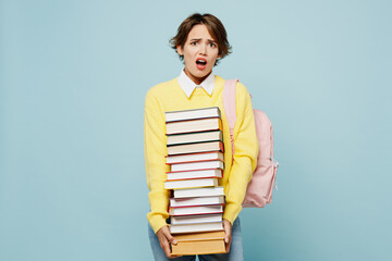 Young shocked dissatisfied indignant woman student wear casual clothes yellow sweater backpack bag hold stack of many books isolated on plain blue background. High school university college concept.