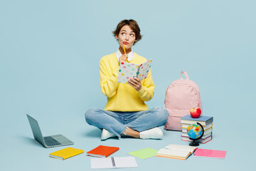 Full body young woman student wear casual clothes sweater sit near backpack bag books write down in exercise book diary notes isolated on plain blue background. High school university college concept.