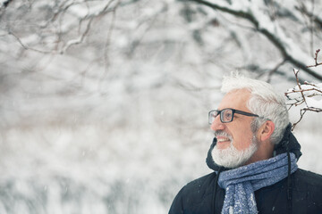 Active grandpa concept. Portrait of handsome mature man in trendy winter clothing and fashionable glasses standing under tree, over snow park background. Close up. Text space.  Outdoor shot