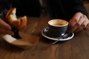 A man's hand holds a round bun and cup of coffee on a wooden table in a cafe. Horizontal indoor...