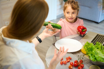 Little girl holding red pepper and giving green cucumber to mother