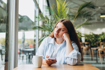 Portrait of sad young redhead woman in a coffee shop looking at smart phone. A lonely unhappy 30s girl siting in a cafe. Bad feelings stress, anxiety, grief, emotions.