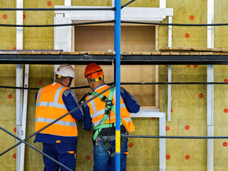 Builders at work. Men at construction site. Two construction workers have their backs to camera. Builders near wall and scaffolding. Installation of insulation on facade. Construction works