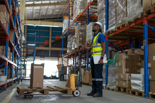 African American man worker pulling pallet truck for move cardboard boxes. Storehouse employee in uniform working and unloading goods in warehouse. Logistics, Distribution Center concept.