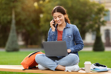 Positive woman student using computer and talking on phone outdoor - obrazy, fototapety, plakaty