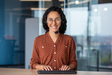 Portrait of young successful Latin American online support worker, woman smiling and looking at camera with headset phone, webcam view, typing on keyboard, inside office at workplace.