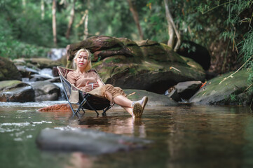 Young woman sitting on a camping chair in the stream and enjoying while holding a coffee cup, Admiring the surrounding natural atmosphere with weak sunlight. Healthy Lifestyle by Nature Therapy.