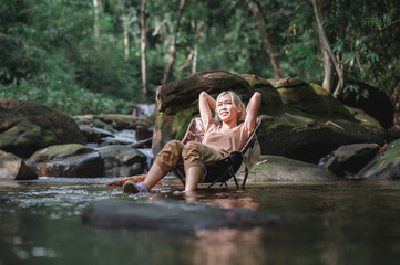 Young woman sitting on a camping chair in the stream and reclining to relax, Admiring the surrounding natural atmosphere with weak sunlight. Healthy Lifestyle by Nature Therapy.