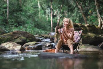 Young woman sitting on a camping chair in the stream and enjoying while holding a coffee cup, Admiring the surrounding natural atmosphere with weak sunlight. Healthy Lifestyle by Nature Therapy.