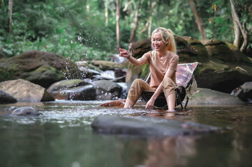 Young woman sitting on a camping chair in the stream and enjoying splashing water, Admiring the surrounding natural atmosphere with weak sunlight. Healthy Lifestyle by Nature Therapy.