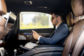 Businessman sits with phone on a backseat of luxury car. Man doing business on the road in prestigious car. Concept of business transportation