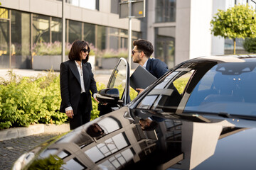 Female chauffeur helps a businessman with laptop to get out of the car, opening a door. Concept of personal driver, luxury taxi or business trips