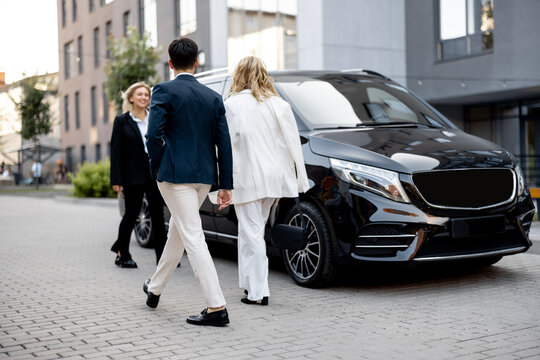 Business people walk to minivan taxi from office building, female driver waits for them nea vehicle. Concept of business trips and transportation service