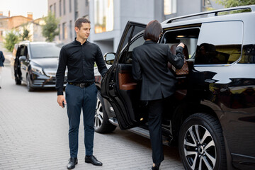 Male driver helps a business lady to get in a car, opening door of a luxury SUV taxi. Business lady with handbag wearing black formal wear. Concept of transportation service - obrazy, fototapety, plakaty