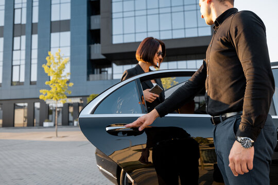 Chauffeur helps an elegant business woman gets in car, opening door of a premium taxi near office building or hotel. Concept of personal driver or business trips