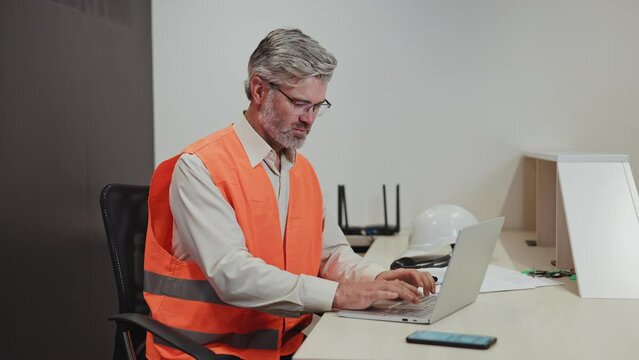 Busy male putting on glasses and starting typing text of new business plan on portable laptop in cabinet. Old repairman sitting at table with hard hat and equipment for comfortable work process.