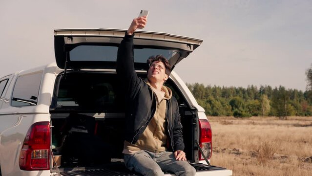 A young guy sits in the open trunk of a car with phone and studies the area during a stop in meadow