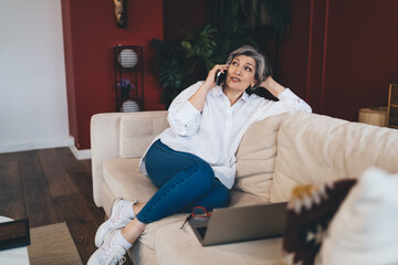 Fototapeta premium Smiling mature woman talking on smartphone on sofa
