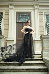 a girl of 30 years old in a sexy, designer, black dress, nylon tights and shoes, posing against the background of the house. Photo shoot, stylish look in a translucent dress. Fashion trends, trends