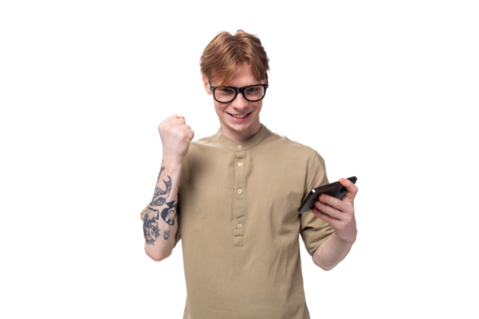 young red-haired european man dressed in a light brown shirt with a tattoo on his arm rejoices holding a phone in his hand