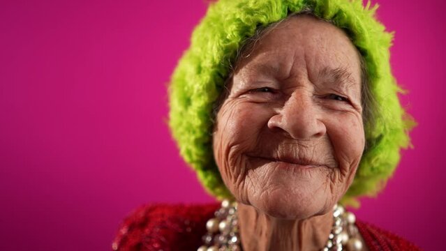 Closeup portrait of smiling fisheye portrait caricature of funny elderly woman smiling looking off camera with green hat isolated on pink background in slow motion.