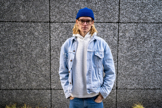 Portrait blond slim guy with long hair, scandinavian androgynous appearance in glasses looking at camera standing on street with hands in pockets. LGBT man gay transgender in denim clothes blue hat. 