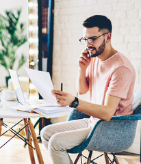 Positive man in optical eyewear working remotely with paper documents calling to colleague