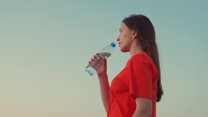 Outdoor sports, girl drinks clear mineral water after training at sunset in sun. Quench your thirst with cool water. Young Woman drinks refreshing water from bottle after training in park in nature