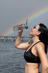 A beautiful and slender girl in sunglasses pours water over herself from a sports bottle against the sky with a rainbow. Sport concept. Close-up.