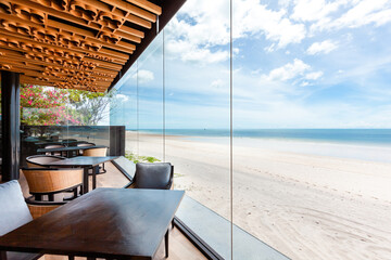 Luxurious beachfront dining area with clear glass walls offering unobstructed ocean views, modern wooden furniture, and vibrant floral accents. Interior design and lifestyle.