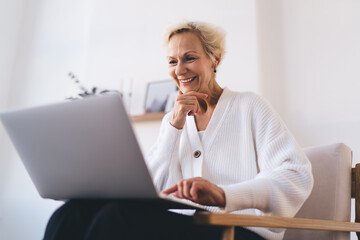 Smiling middle aged woman working on laptop and sitting on armchair