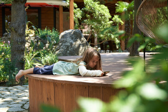 A three - year - old boy with a broken arm plays games on a mobile phone , lying on his stomach , among the greenery in the resort.