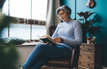 Focused mature woman reading book in room