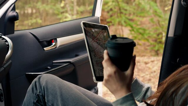Close-up shot of a girl sitting in a car with a cup of tea and plotting a route through the forest using a map on tablet