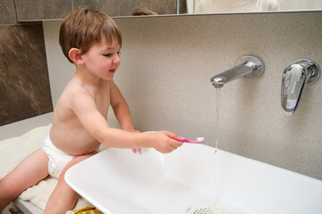 The child washes their toothbrush in the bathroom sink after brushing their teeth. Kid aged two...