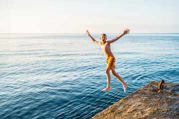 Healthy boy jumps into tranquil ocean water from stone pier in sunny morning. Sportive child dives...